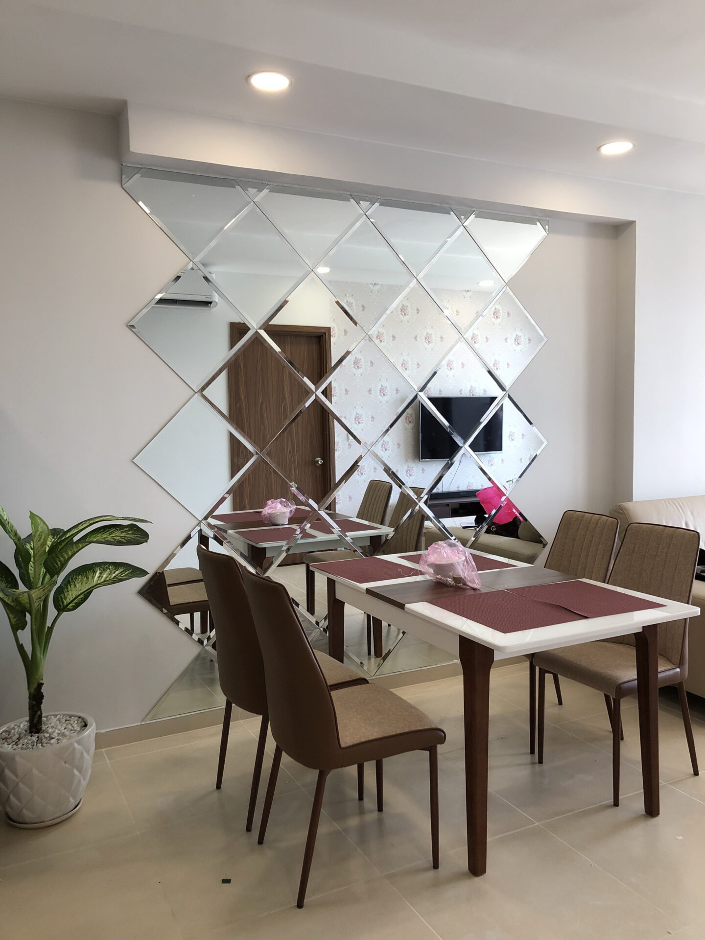Kính trang trí phòng ăn, vách kính ốp tường nghệ thuật hình thoi, bàn ăn gỗ, ghế bọc nệm, và chậu cây xanh.